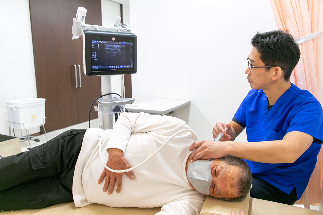 医療従事者が診察台に横たわる高齢の患者の首元を、超音波画像を映すモニターを見ながらエコー検査している様子